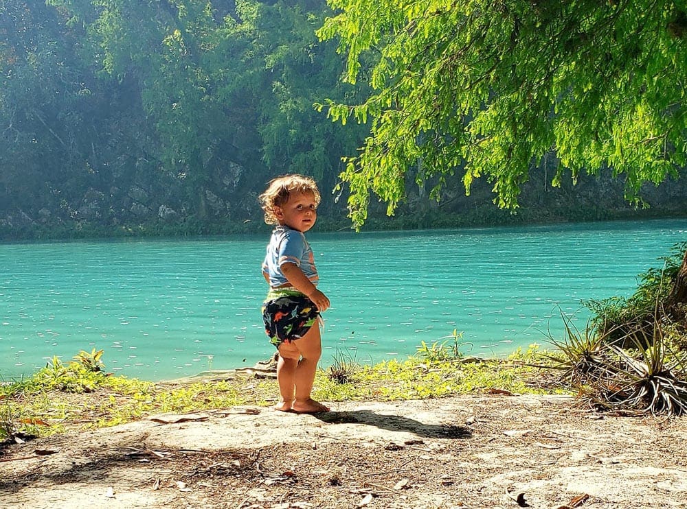 A toddler looks back at the camera with an expansive view of clear blue waters and tropical foliage behind her.