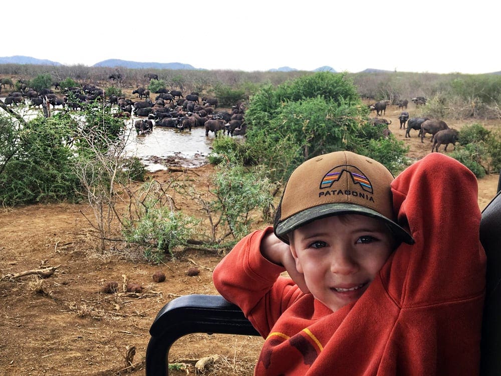 A young boy smiles at the camera, with a watering hole and hundreds of water buffalo behind him while on safari in Africa.