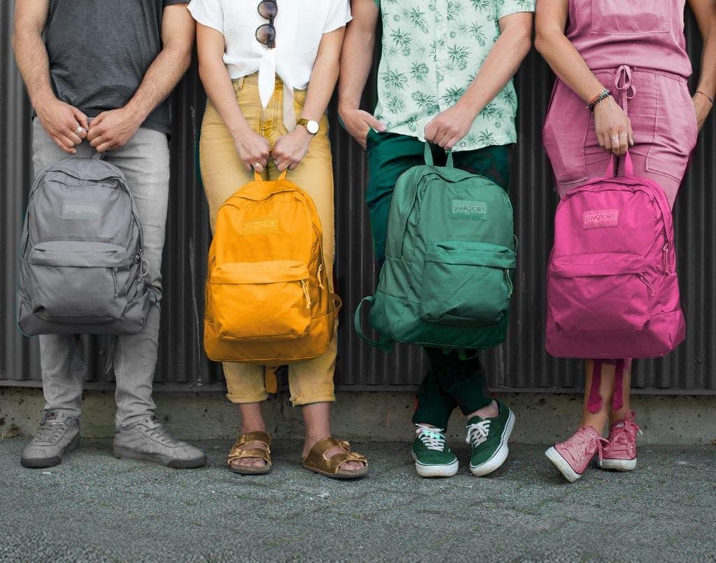 the bottom halves of four people holding grey, orange, green, and pink JanSport backpacks, each has color-coordinated clothing. 