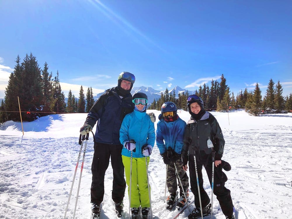 A family of four wearing snow gear stands on skis in the snow at Telluride with trees and snow behind them.