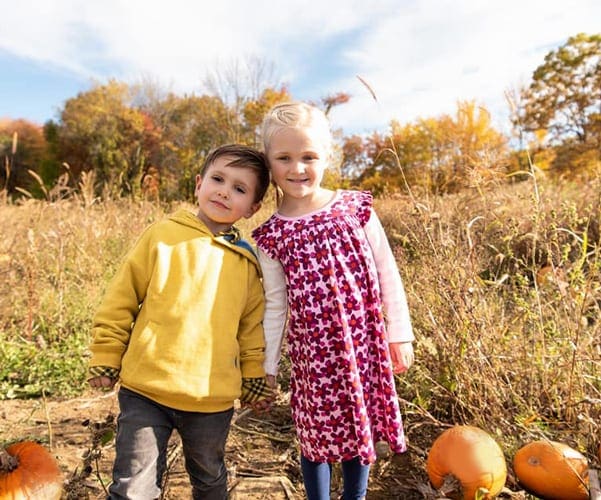 Two kids stand shoulder to shoulder in a pumpkin patch.