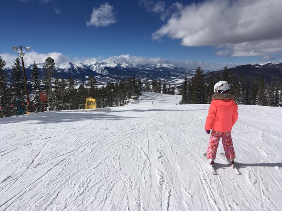 A young child wearing a pink snowsuit and white helmet looks down the slope at Ketstone.