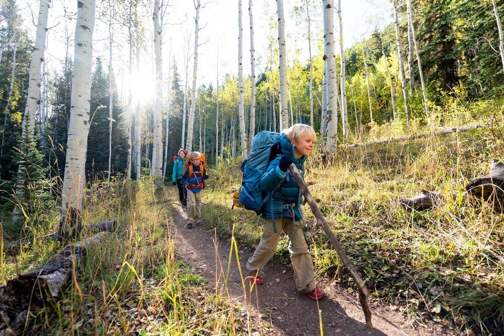 A young boy wearing an Osprey Pack leads the trail with a walking stick in his hand, two other hikers follow him.