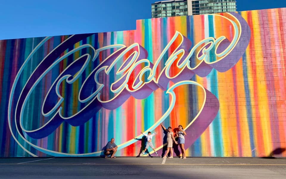Three kids and their dad stand excited in front of a colorful mural in Honolulu reading "Aloha".