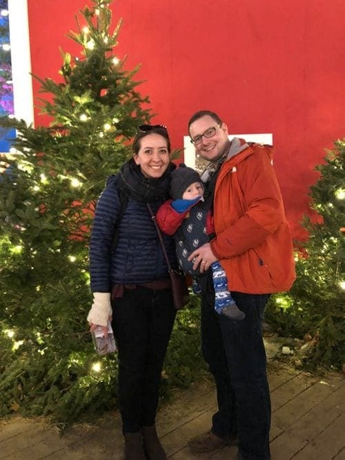 Two parents and their baby stand in front of several lit-up Christmas trees in Freeport, Maine.