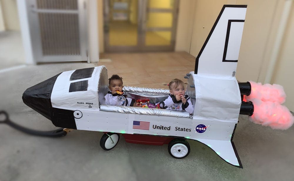 Two infants dressed as astronauts ride in a wagon converted to look like a United States NASA rocket, one of the best travel themed costumes for kids.