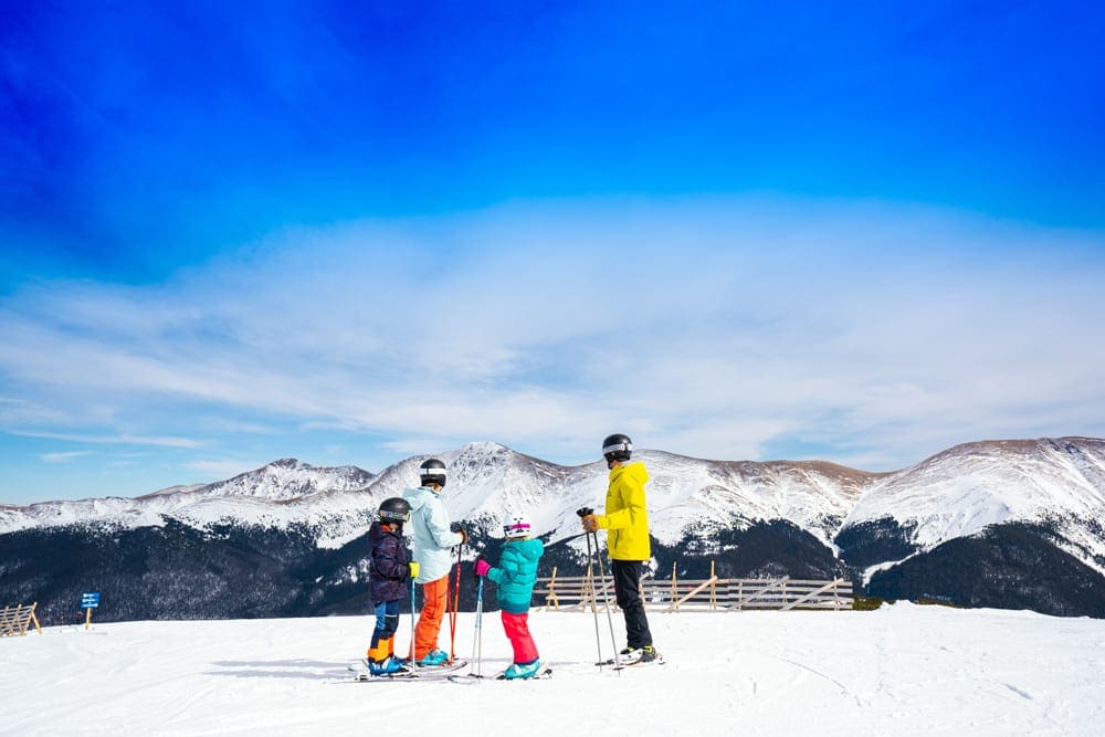 A family of four wearing brighting colored snowgear stands in skis at Winter Park on a clear blue day.