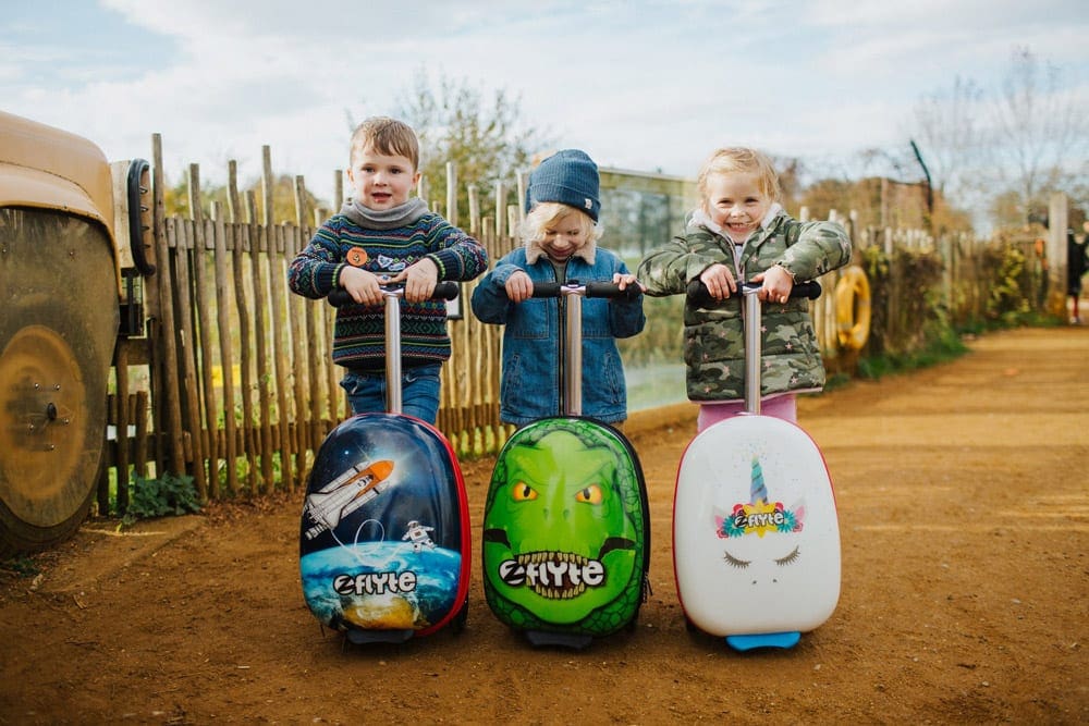 Three kids stand proudly with their Zinc Flyte luggage, featuring a dinosaur, a uninorn, and a space-inspired suitcase. Zinc Flyte is one of the best travel luggage for kids.