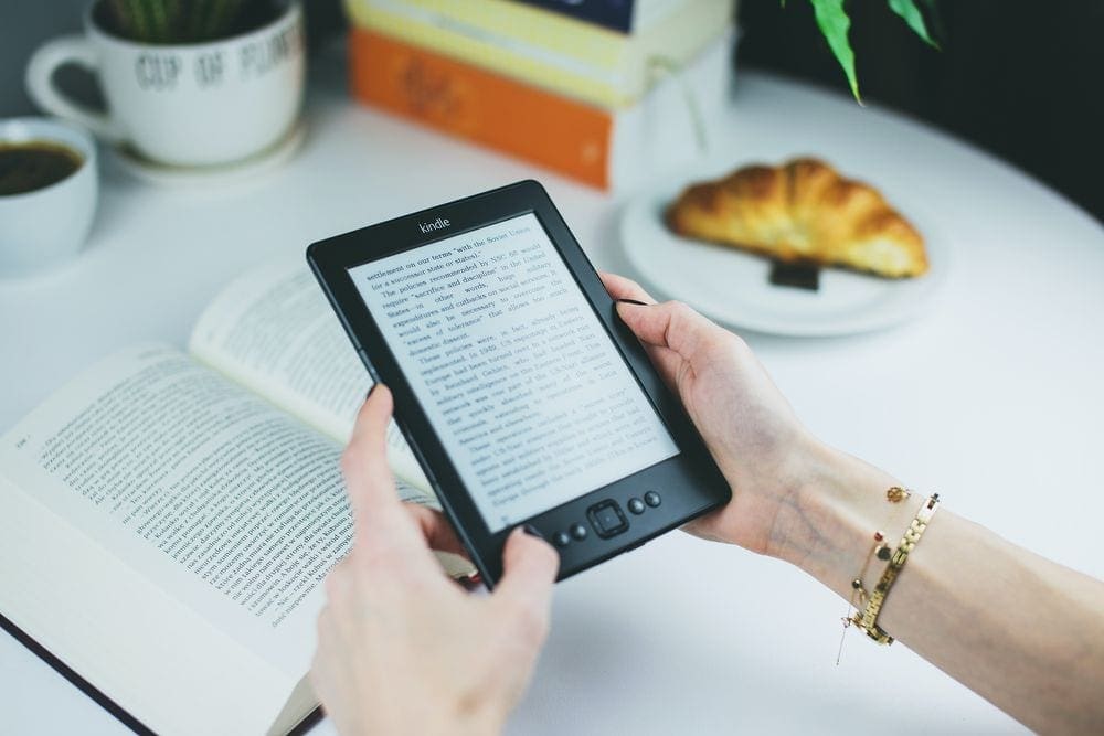 A woman's hands hold a kindle over a book, with a croissant and coffee nearby on the table.