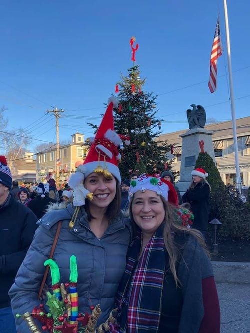 Two women wearing festive Christmas hats stand in front of a city Christmas tree in Kennebunkport.
