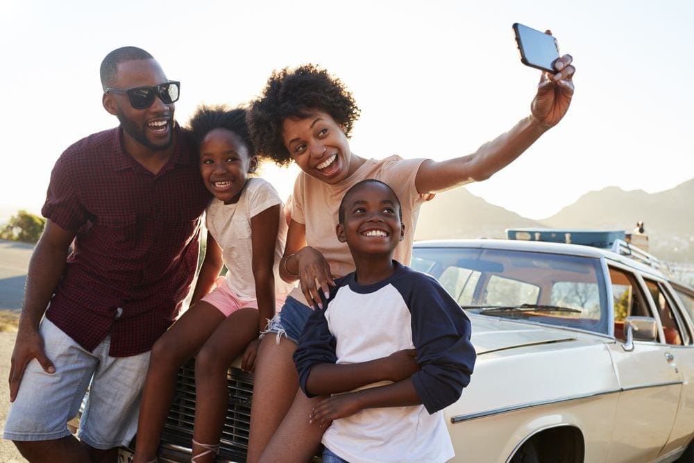 A black mom holds out her hand with an iPhone to take a selfie of her family near their car, knowing the where to get your rental car is just one of the things on our list of travel resources for families.