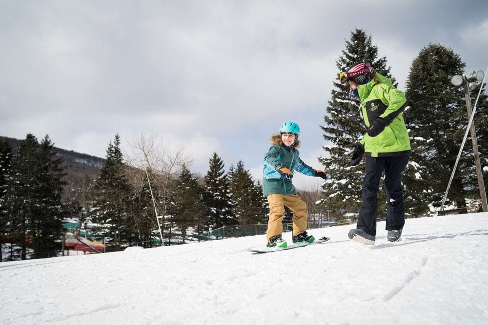 A young child slowly goes down a run on a snowboard, with their ski instructor coaching them each step of the way at Camelback Resort, one of the best ski resorts near NYC for families.