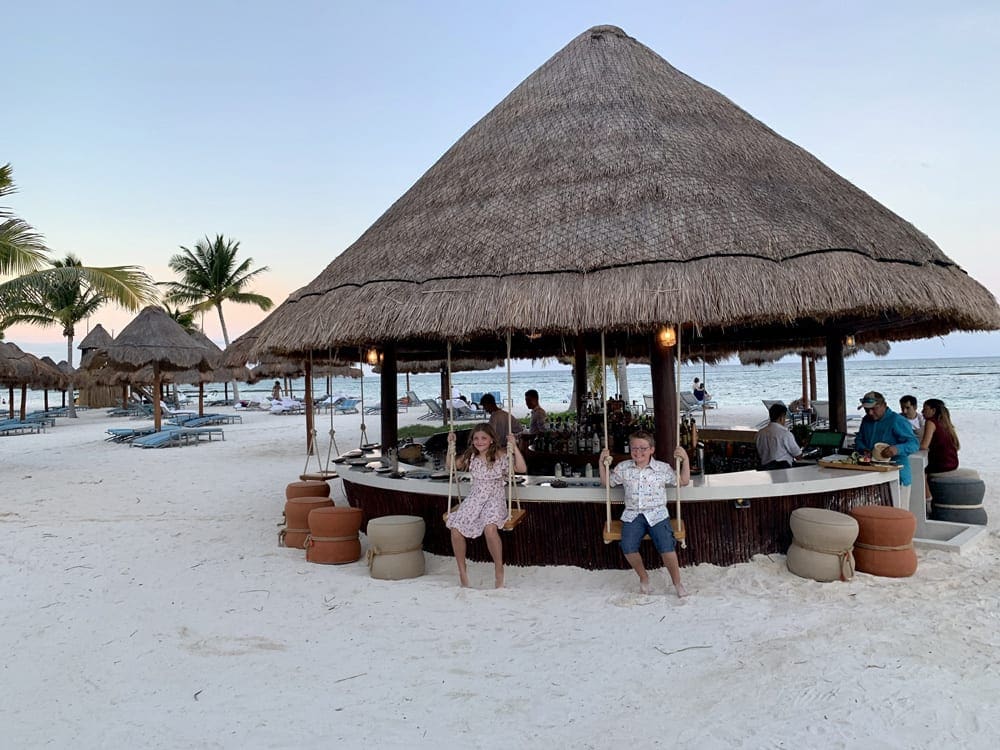 Two kids sit on swings, at a juice bar on-site at Fairmont Mayakoba.