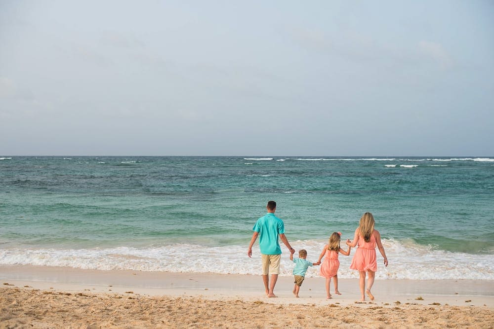 A family of four, wearing color coordinated outfits in blue and pink, walks on the beach toward the water in the Dominican Republic, one of the best Caribbean islands for families.