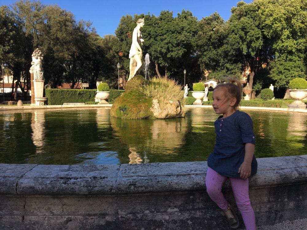 A young girl sits on the edge of a pond within the Borghese Gardens.