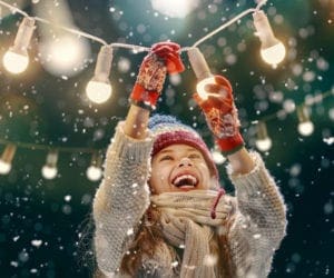 A young girl with a huge smile reaches up to touch twinkling lights on a winter day.