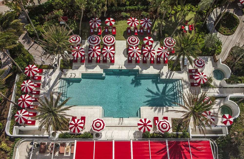 An aeiral view of a large turquois pool at Faena Hotel Miami Beach, surrounded by the iconic candy red chairs and umbrellas.