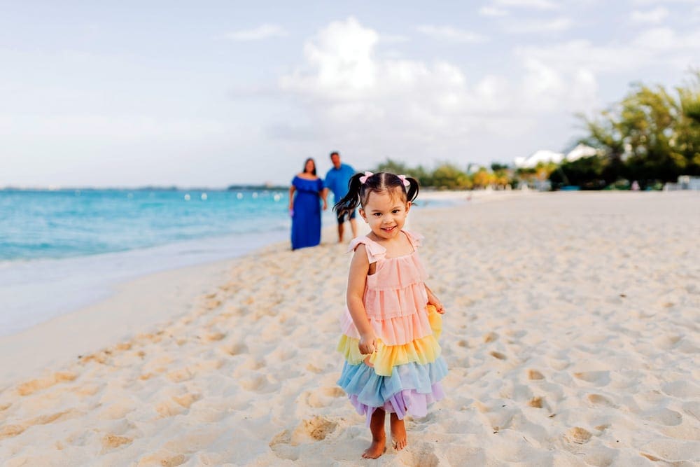 A small girl wearing a colorful dress smiles while her parents look on in the background on a beach in the Cayman Islands, one of the best hot places to visit in December for families.