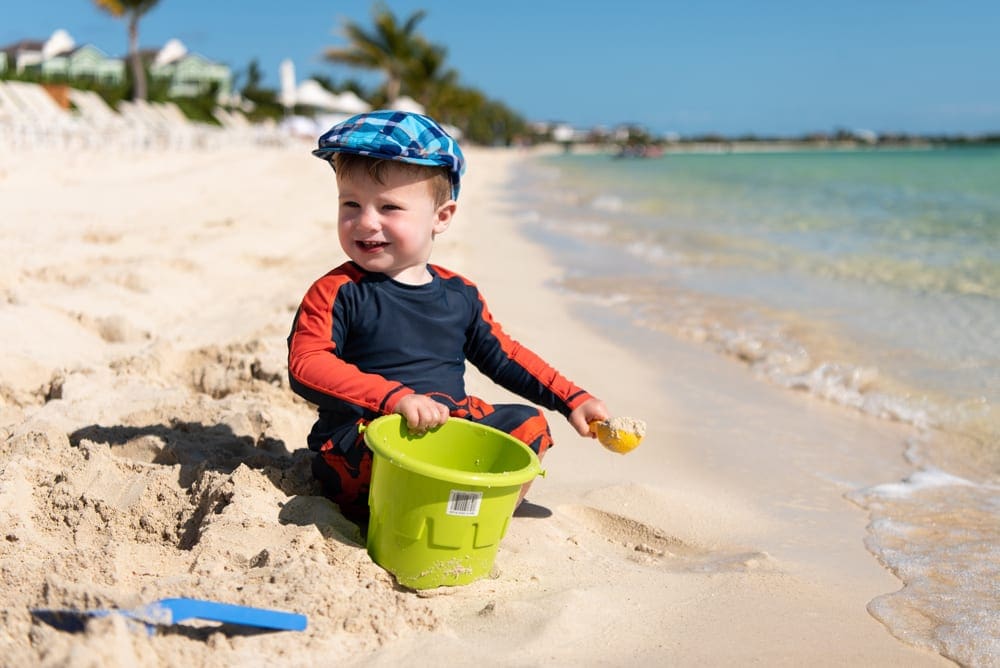 A young boy wearing a cap sits smiling in the sand on the beach.