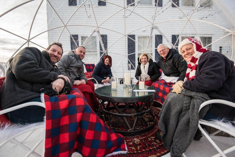 A family of six, wearing winter clothes and blankets, sits around an outdoor table within a globe at the Kennebunkport Inn.