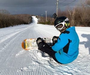 A young boy sits in the snow on a slope at Blue Mountain while resting his board.