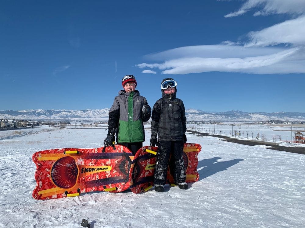 Two boys, well dressed for sledding in the winter, stand near their red and yellow sled at Fraser Hill.