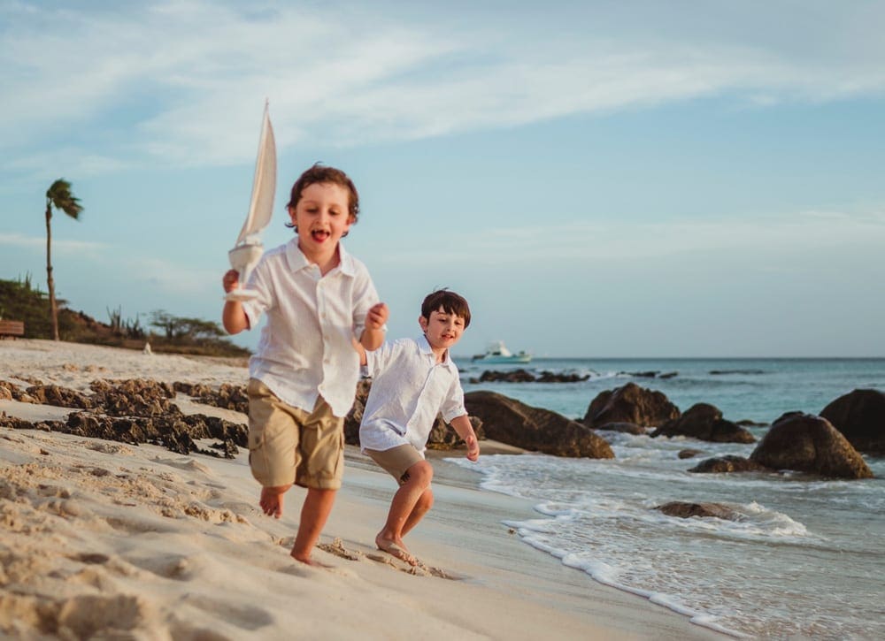 Two boys race down a beach in Aruba, while one carrys a toy sail boat.