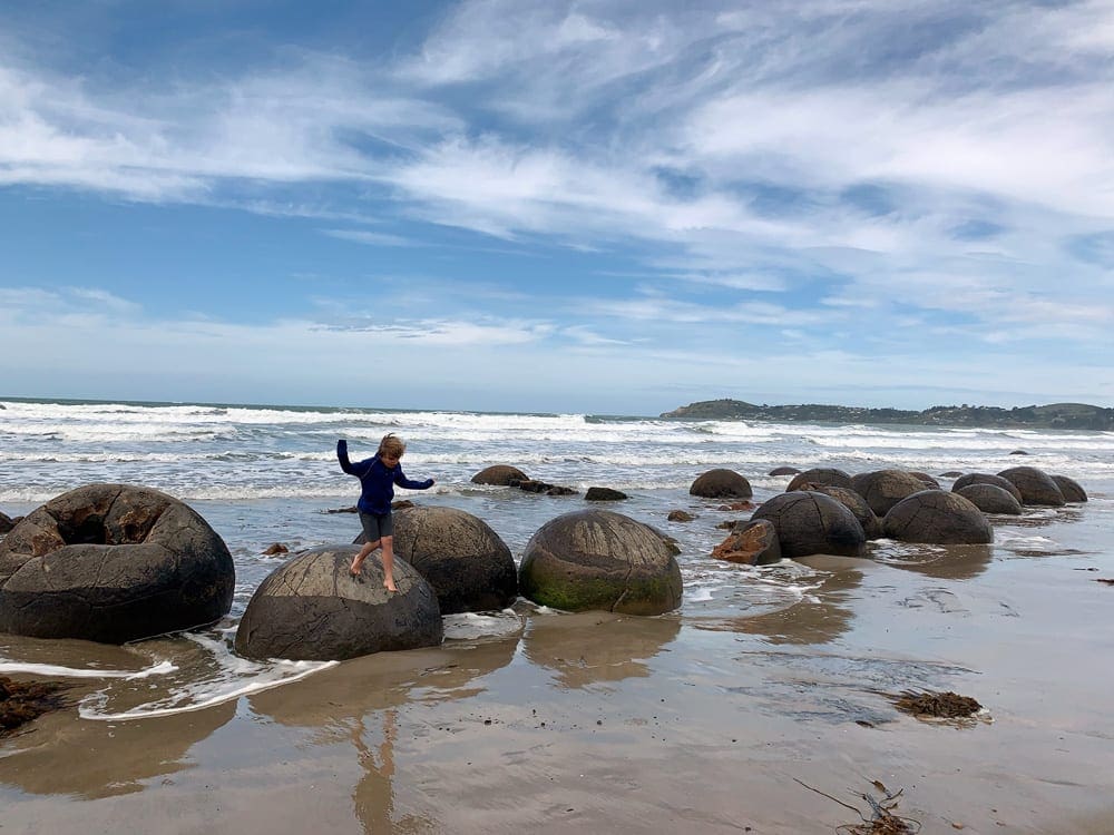 A young boy bounds from bolder to bolder on a beach in New Zealand, one of the best places for a December vacation with kids.