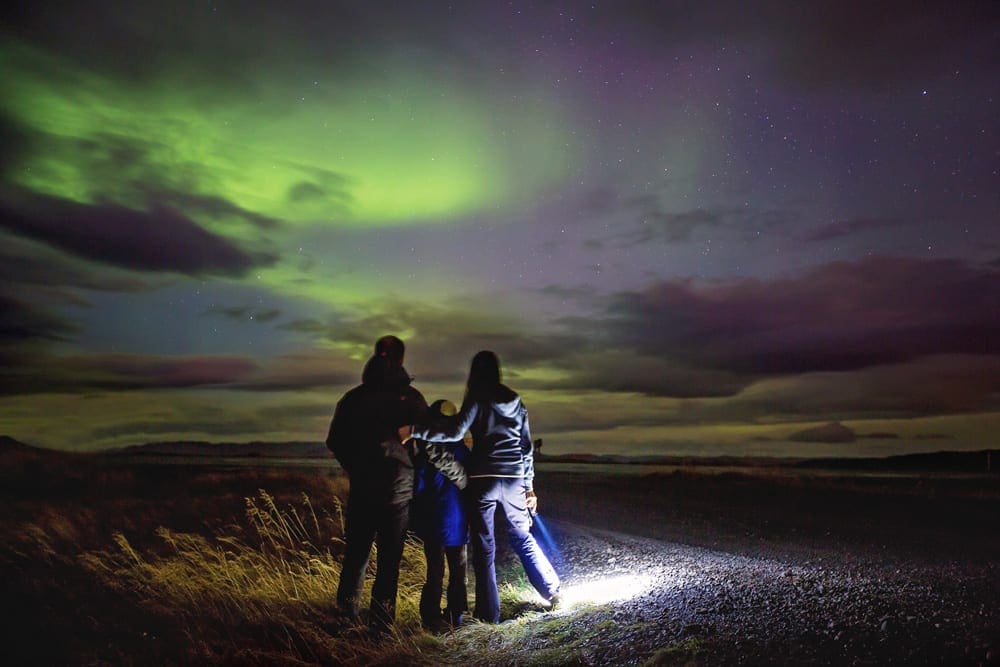 A family of three stands looking off onto stunning northern lights in hues of green and blue.