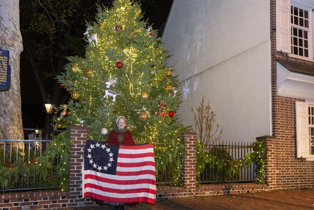 A woman holds a colonial flag outside of Betsy Ross's House in Philadelphia, a large Christmas tree stands behind her.