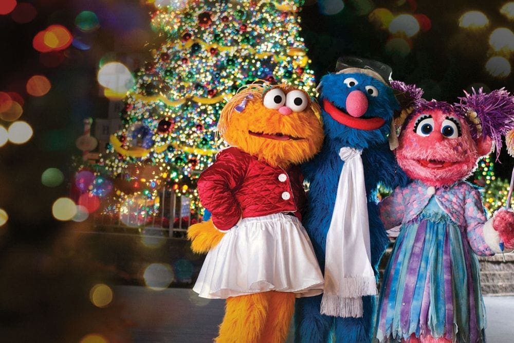 Three Seasame Street charachters stand in front of a large Christmas tree at Sesame Place.