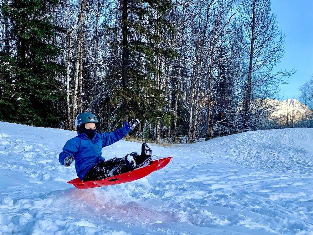 A young boy wearing a blue helmet and a blue coat sits on a red sled airborne, as it goes over a small snow mound. Sleds are some of the best outdoor gifts for families.