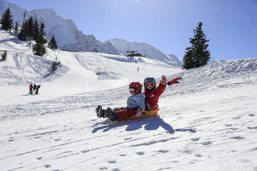 A young child waves while riding on a yellow sled behind another child. Sledding is , one of the best things to do in Chamonix with kids in Winter.