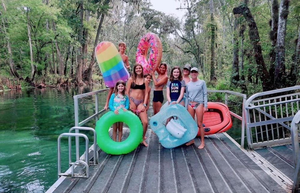 Eight people, including some young kids, hold several colorful inflatable tubes, surrounded by a turqoius river.