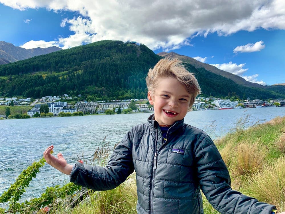 A young boy holds his arms out wide smiling with a sweeping view of Queenstown behind him.