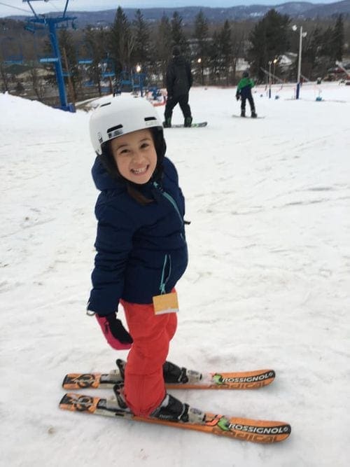 A young girl on skis, wearing a helmet, smiles brightly at the top of the bunny hill.