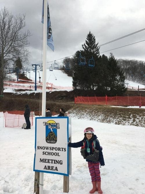 A young girl wearing snow gear and a helmet leans agains a sign that reads "Thunder Ridge Snowsports School Meeting Area", one of the best ski resorts near NYC for families.