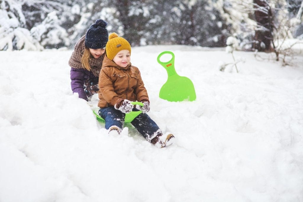 A young girl pushes her little brother down a small hill on a green sled.