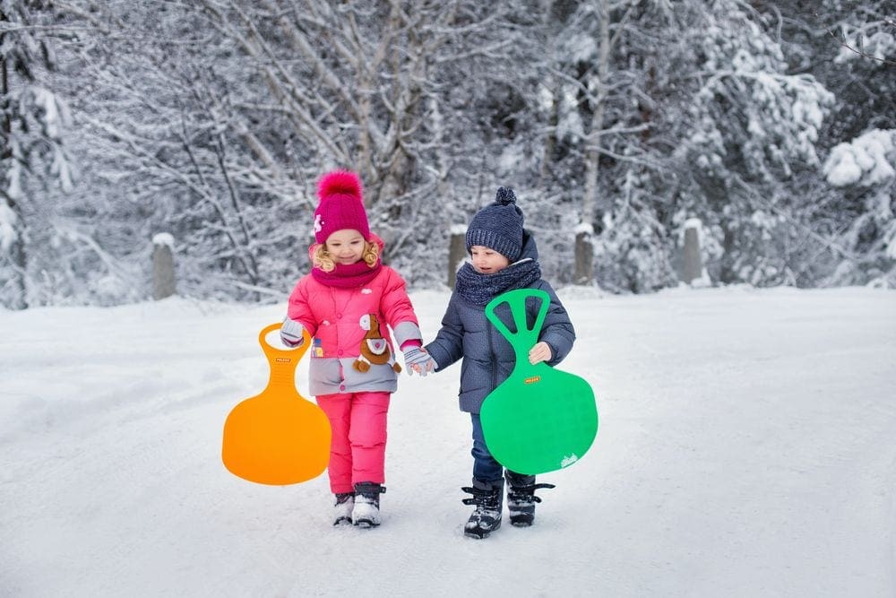 A young boy wearing gray and young girl wearing pink hold hands as they walk in the snow with their sleds.