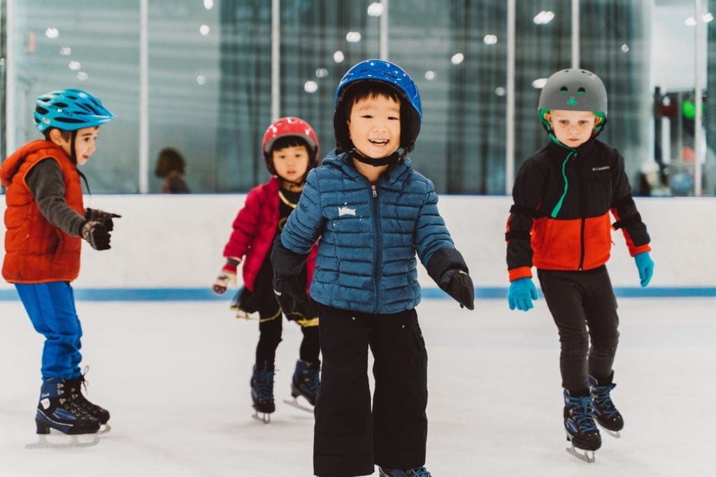 Several young boys wearing winter gear and helmets skate along the rink at Sky Rink at Chelsea Piers, one of the best places to go ice skating in NYC with kids.