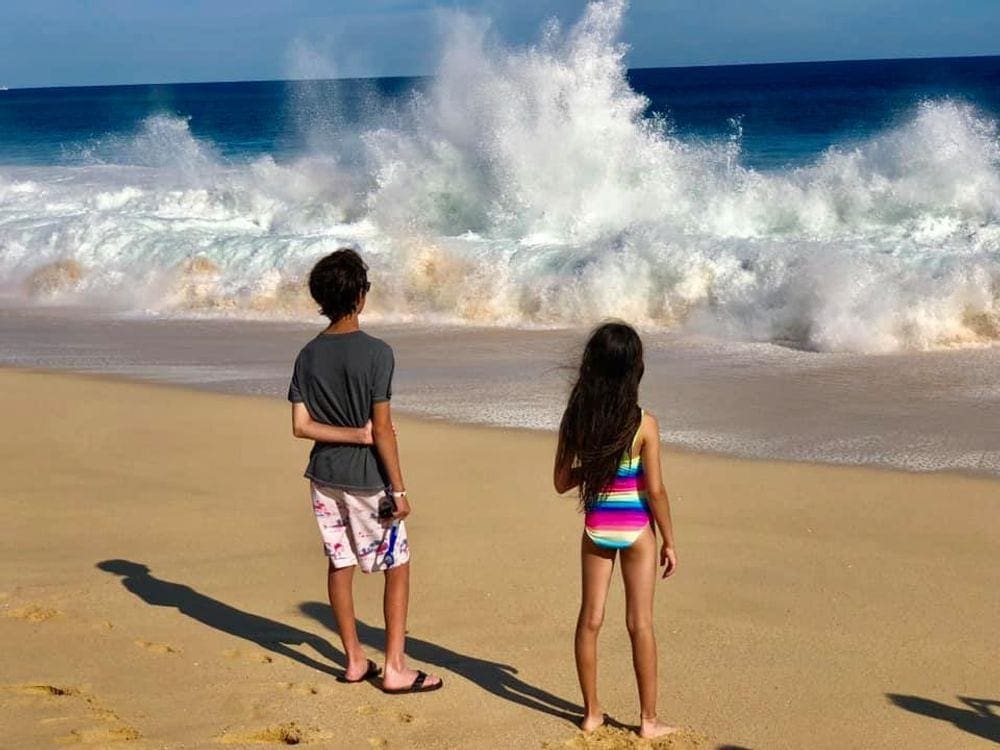 Two young kids stand on the beach in Los Cabos, while looking at crashing waves, one of the best hot places to visit in December for families.