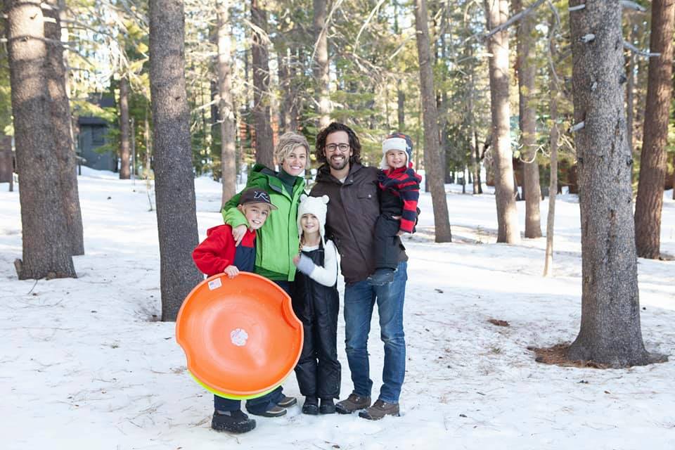 A family of five stands in the snow, wearing full snow gear. One child is holding an orange sled.