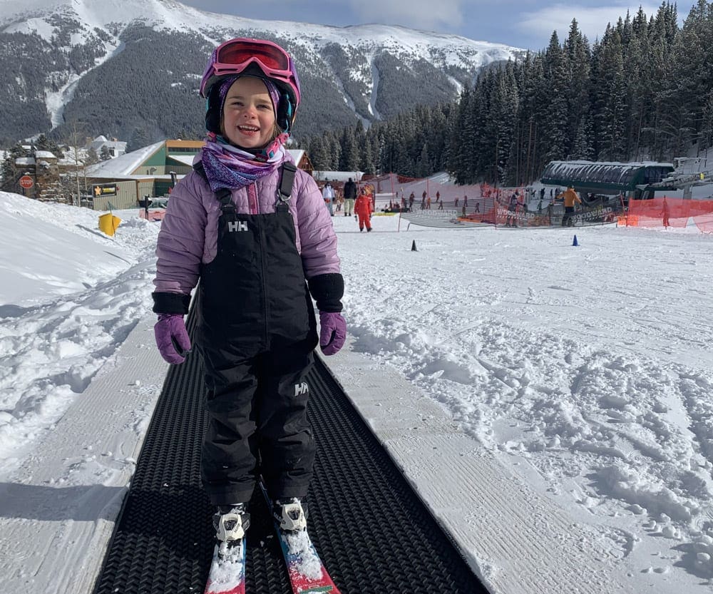 A young girl wearing a full snow suit and helmet stands on skis on a conveyor, all of these items are things to remember in our Ultimate Ski Trip Packing List For Families This Winter.