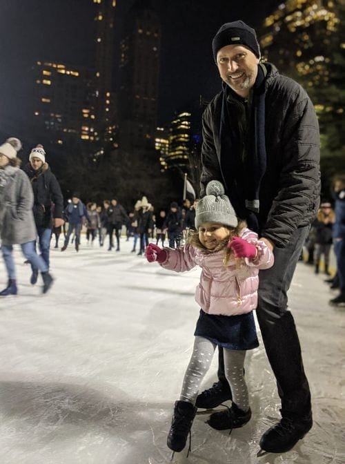 A father holds his young daughter up on skates while ice skating in Central Park.