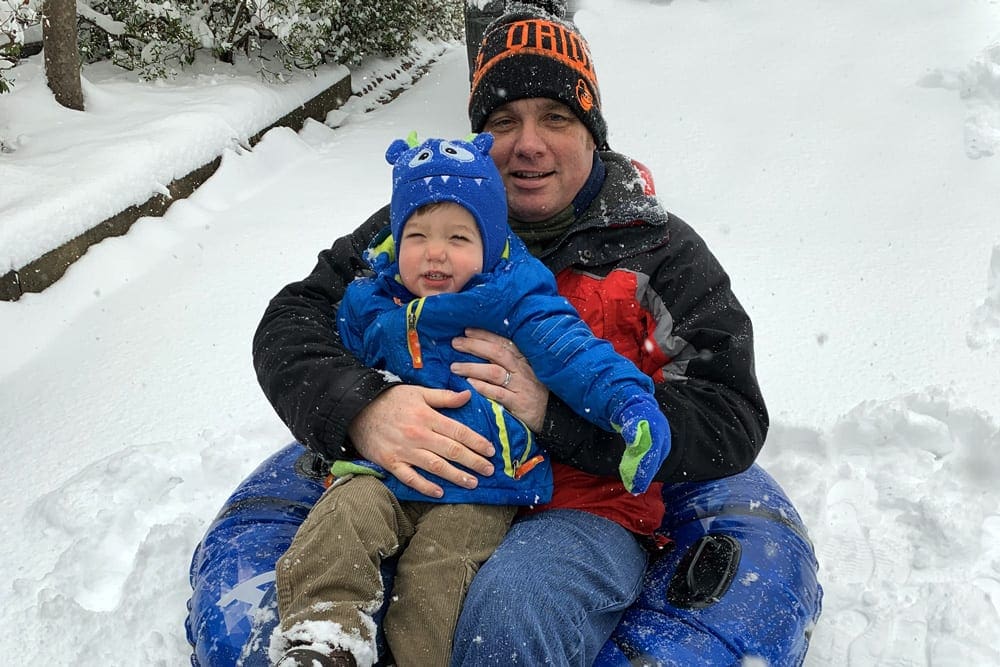 A dad and his young son wear full snow gear while sitting on a blue snow tube.
