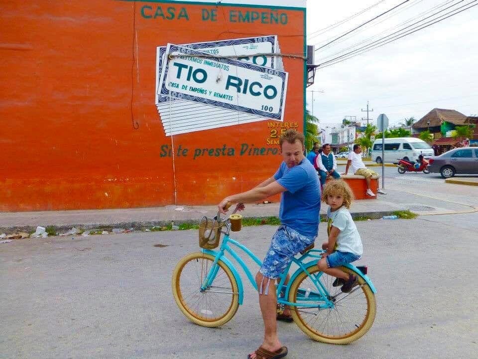 A young boy sits on the back of his father's turquoise bike next to an orange building in Playa del Carmen.