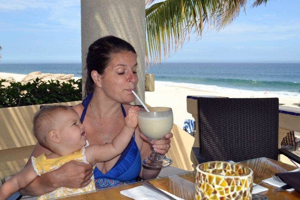 A mom drinks from a tropical drink in Los Cabos as her young infant sits on her lap and reaches out.