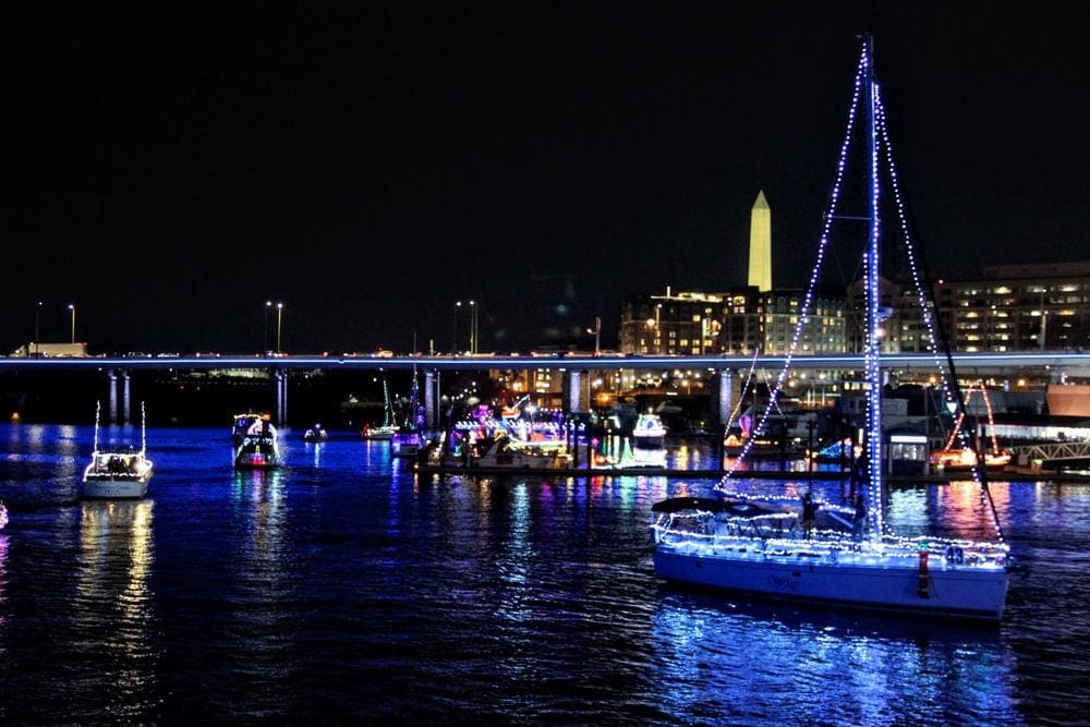 Several ships are lit with holiday lights at The Wharf in Washington DC, one of the best places to celebrate the holidays in DC with kids, with the Washington Monument shown in the distance.