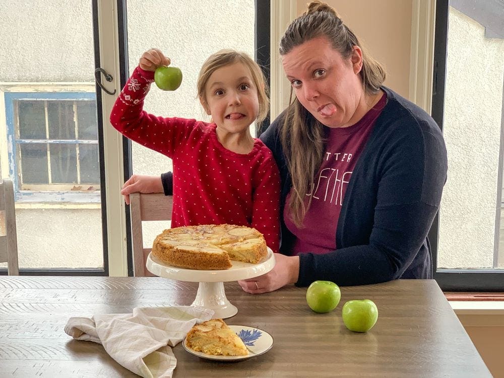 A young girl and her mom make silly faces as they sit next to a plated apple shartlota.