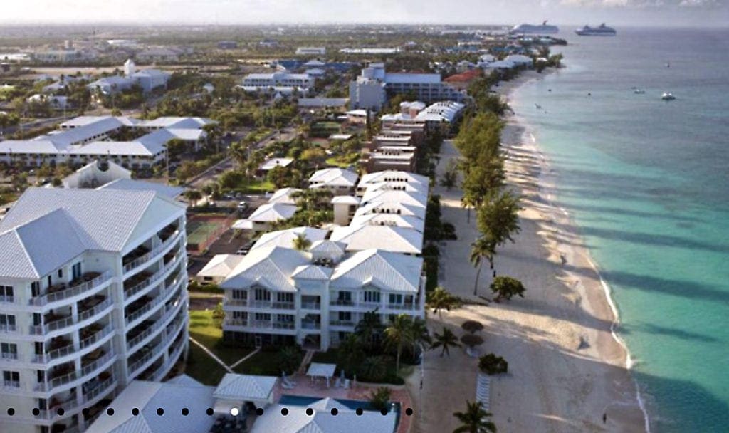 An aerial view of Caribbean Club Hotel Grand Cayman, one of the best family resorts in Grand Cayman, featuring an expansive beach and large hotel buildings.